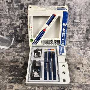 May include: A Staedtler Marsmatic technical pen set. The set includes a box with a blue and red pen, and a tray with multiple pens and accessories. The box has the text "marsmatic" and "STAEDTLER".