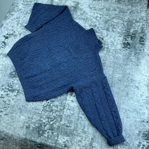 May include: A dark blue speckled knit sweater featuring a high collar with a zipper closure, long sleeves with vertical cable knit detailing, and a ribbed hem. The garment is crafted with a textured weave and includes a small chest pocket.