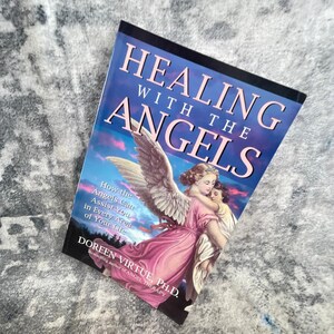 May include: A paperback book titled Healing with the Angels by Doreen Virtue, Ph.D. The cover features an illustration of an angel with large wings embracing a child against a blue and purple sunset sky with clouds and a dark forest silhouette.