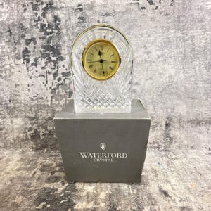 May include: A clear crystal clock with a gold-colored clock face and hands. The clock is encased in a decorative, cut crystal frame. The clock is sitting on top of a gray box with the words "WATERFORD CRYSTAL" printed on it.