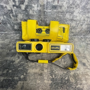May include: A bright yellow Minolta Weathermatic 35mm film camera with a matching yellow carrying case. The camera features a rectangular body with a black lens, a flash unit, and a black strap. The case displays a line drawing of the camera.