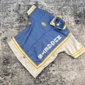 May include: A light blue and white jacket with the word "NUGGETS" in white block letters. The jacket has yellow stripes and a gold emblem. The jacket is a short-sleeved style.