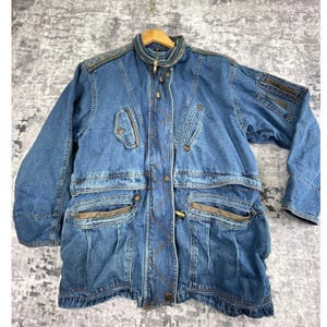 May include: A vintage denim jacket with a blue wash, featuring multiple pockets, a zip-up front, and decorative stitching. The jacket has a collared neckline and is hanging on a wooden hanger. The jacket has a unique design with brown accents.