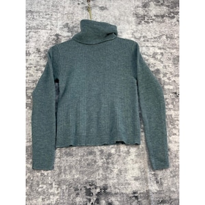 May include: A teal-colored turtleneck sweater with ribbed knit detailing. The long-sleeved sweater is hanging against a textured background. The collar is folded over, and the sweater is made of a soft material.