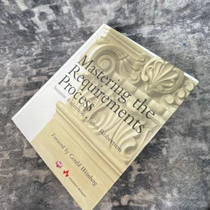 May include: A hardcover book titled Mastering the Requirements Process by Suzanne Robertson and James Robertson, featuring a cream-colored cover with a large, detailed illustration of a classical architectural column capital in beige tones.