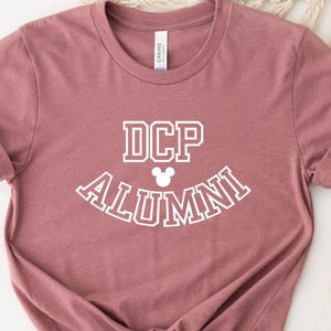 May include: A dusty rose-colored t-shirt with the text "DCP ALUMNI" in white, arched across the front. A small Mickey Mouse head graphic is between the letters "DCP" and "ALUMNI". The shirt has rolled-up sleeves and a knotted hem.