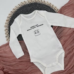 Könnte beinhalten: Weißer Baby-Body mit langen Ärmeln. Auf dem Body steht der Text "ERROR 404 SCHLAF NICHT GEFUNDEN" und ein Cartoon-Gesicht. Er liegt auf einer grauen Webmatte und einem rosa Stoff.