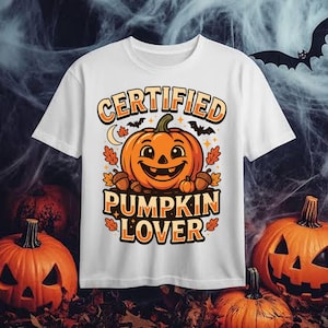 May include: A white short-sleeve t-shirt featuring a graphic of a smiling orange jack-o'-lantern surrounded by autumn leaves, acorns, and bats. The text "CERTIFIED PUMPKIN LOVER" is printed in bold orange and black letters above and below the pumpkin.