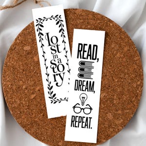 Puede incluir: Dos marcadores blancos con texto y diseños en negro. Un marcador dice "Lost in a Story" dentro de un borde de hojas. El otro dice "Read, Dream, Repeat" con gráficos de libros, bombillas y gafas. Los marcadores están sobre una superficie de corcho.
