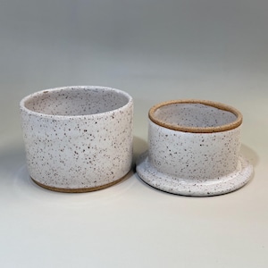 Pu&ograve; includere: Una burriera in ceramica con smalto bianco maculato. Il piatto &egrave; composto da una base cilindrica e un coperchio con la parte superiore arrotondata e un piccolo bordo. Il bordo e la base hanno una finitura naturale non smaltata.
