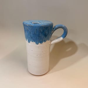 May include: A handmade ceramic mug with a unique design. The top portion is a vibrant blue, with a dripped glaze effect, while the lower part is white. It features a curved handle and a hole in the top, likely for a specific purpose.