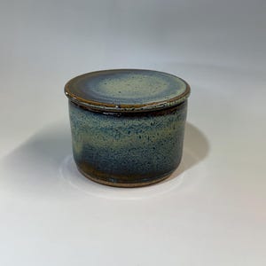 Pu&ograve; includere: Un vaso in ceramica fatto a mano con coperchio. Il vaso &egrave; cilindrico con una glassa blu-verde screziata e un bordo marrone scuro. Il coperchio &egrave; rotondo, in tinta con lo schema cromatico del vaso. La ceramica ha un aspetto rustico e artigianale.