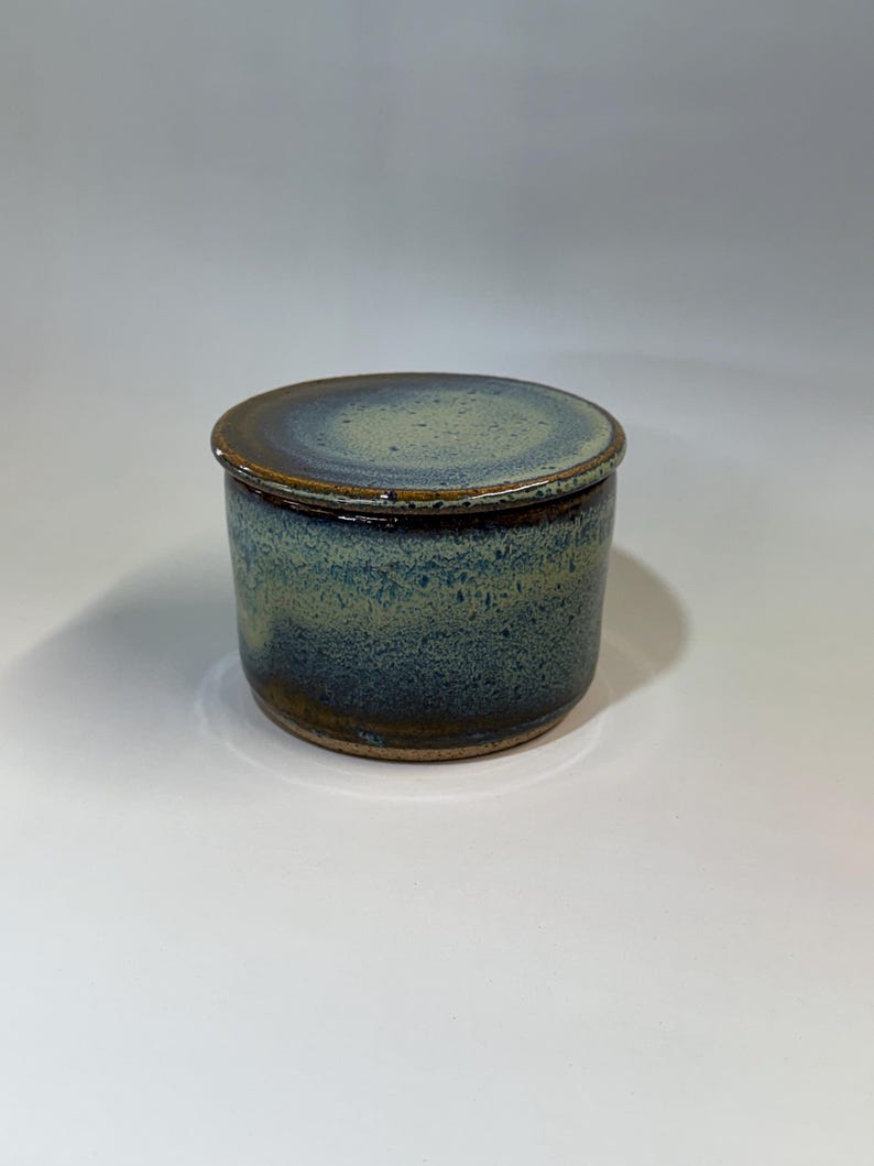 Pu&ograve; includere: Un vaso in ceramica fatto a mano con coperchio. Il vaso &egrave; cilindrico con una glassa blu-verde screziata e un bordo marrone scuro. Il coperchio &egrave; rotondo, in tinta con lo schema cromatico del vaso. La ceramica ha un aspetto rustico e artigianale.