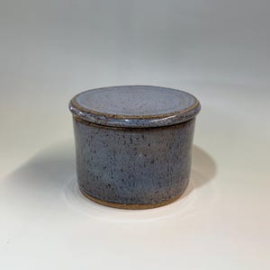 Pu&ograve; includere: Un burriera rotonda in ceramica con coperchio. Il piatto e il coperchio sono di colore blu-grigio maculato con bordo e base marroni. La burriera &egrave; appoggiata su una superficie bianca.