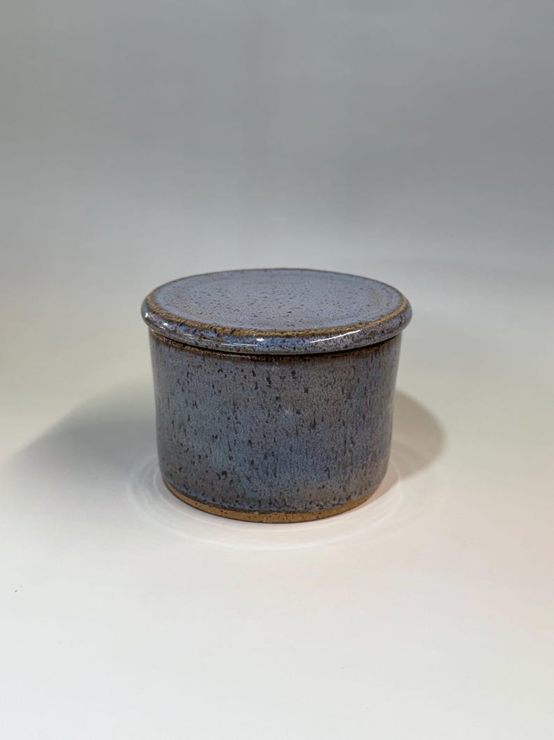 Pu&ograve; includere: Un burriera rotonda in ceramica con coperchio. Il piatto e il coperchio sono di colore blu-grigio maculato con bordo e base marroni. La burriera &egrave; appoggiata su una superficie bianca.