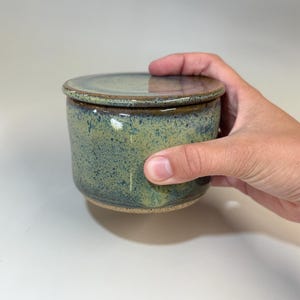 Pu&ograve; includere: Una burriera in ceramica fatta a mano con coperchio. Il piatto &egrave; cilindrico con una glassa verde-blu screziata e un bordo marrone. Il coperchio &egrave; piatto e si abbina alla combinazione di colori del piatto. Una mano tiene la burriera.