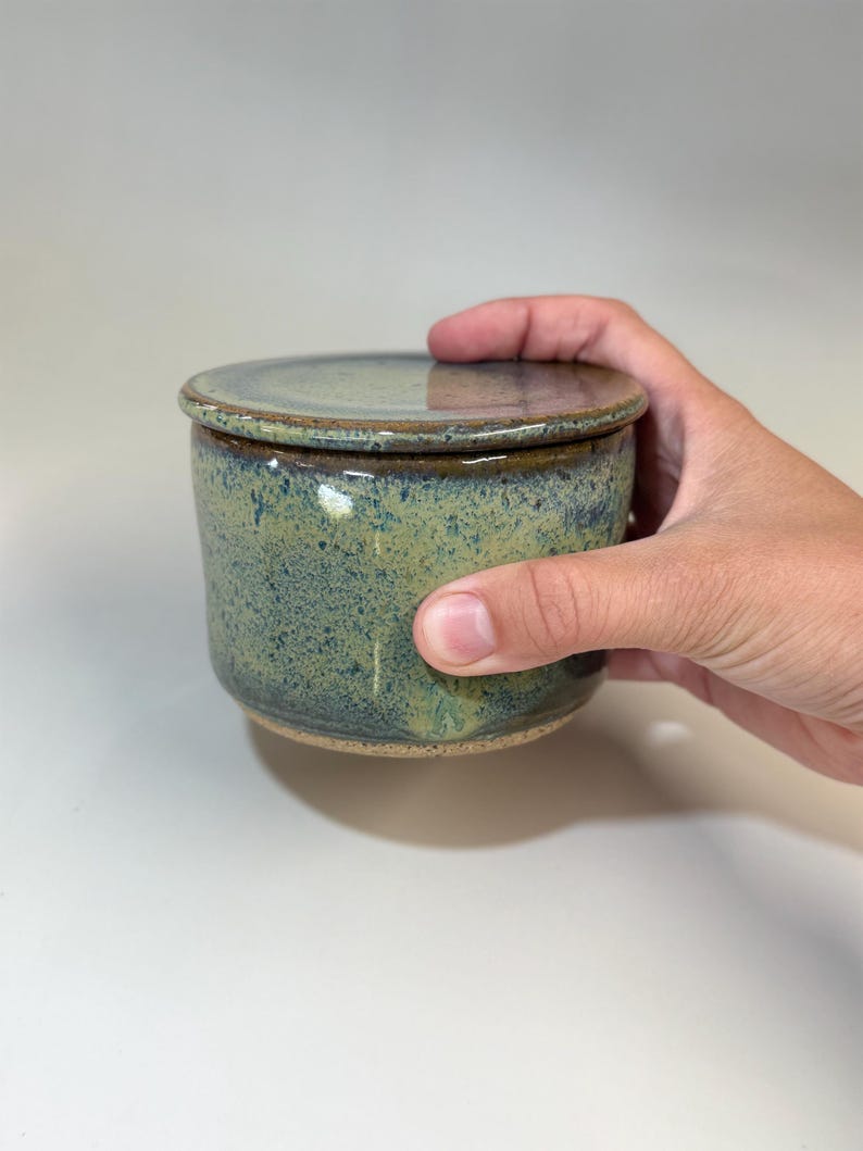 Pu&ograve; includere: Una burriera in ceramica fatta a mano con coperchio. Il piatto &egrave; cilindrico con una glassa verde-blu screziata e un bordo marrone. Il coperchio &egrave; piatto e si abbina alla combinazione di colori del piatto. Una mano tiene la burriera.