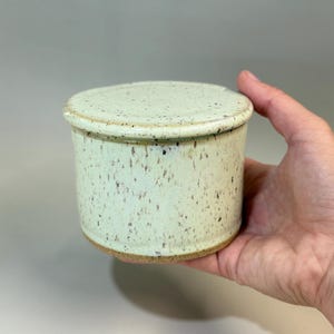 Pu&ograve; includere: Una burriera in ceramica con coperchio. Il piatto cilindrico ha una glassa maculata verde chiaro e un bordo marrone. Il coperchio rotondo corrisponde al piatto. Il piatto &egrave; tenuto in una mano.