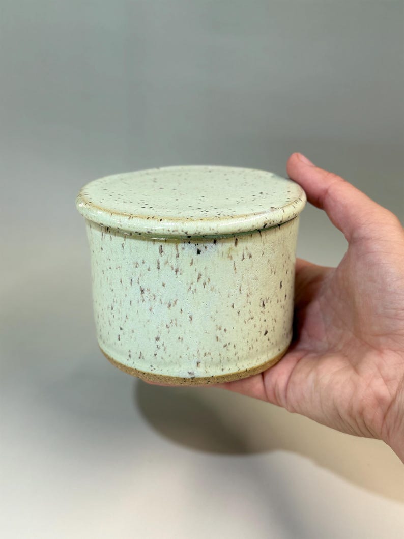 Pu&ograve; includere: Una burriera in ceramica con coperchio. Il piatto cilindrico ha una glassa maculata verde chiaro e un bordo marrone. Il coperchio rotondo corrisponde al piatto. Il piatto &egrave; tenuto in una mano.