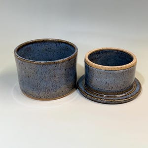Pu&ograve; includere: Burriera in ceramica in due pezzi con smalto blu screziato. Il piatto &egrave; composto da una base cilindrica e un coperchio abbinato con la parte superiore arrotondata. Il bordo e i bordi hanno una tonalit&agrave; marrone pi&ugrave; scura, aggiungendo contrasto. Fatto a mano.