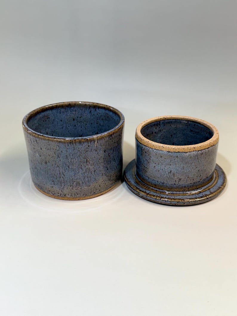 Pu&ograve; includere: Burriera in ceramica in due pezzi con smalto blu screziato. Il piatto &egrave; composto da una base cilindrica e un coperchio abbinato con la parte superiore arrotondata. Il bordo e i bordi hanno una tonalit&agrave; marrone pi&ugrave; scura, aggiungendo contrasto. Fatto a mano.