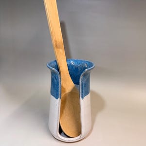 May include: A handmade ceramic utensil holder with a blue rim and white body, holding a wooden spoon. The holder is approximately 6 inches tall and 4 inches wide, perfect for kitchen organization.