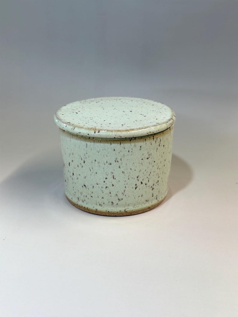 Pu&ograve; includere: Una burriera in ceramica con coperchio. Il piatto cilindrico ha una glassa maculata verde chiaro e un bordo marrone pi&ugrave; scuro. Il coperchio rotondo si adatta perfettamente. Un articolo di ceramica fatto a mano.