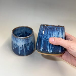 May include: Two ceramic tumblers featuring a deep blue base glaze with a white drip effect cascading from the rim. The cups have a rounded, handleless shape with a smooth, glossy finish and a visible brown clay rim at the top and base edges.