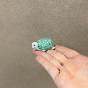 May include: A small, ceramic turtle figurine with a turquoise shell and white head and feet. The turtle has a smiling face and is a decorative item.
