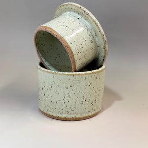 Pu&ograve; includere: Un burriera in ceramica in due pezzi. La base &egrave; un contenitore cilindrico e il coperchio &egrave; un piatto poco profondo. Entrambi i pezzi sono verde chiaro con macchie marroni e un bordo marrone. Il coperchio poggia sulla base.