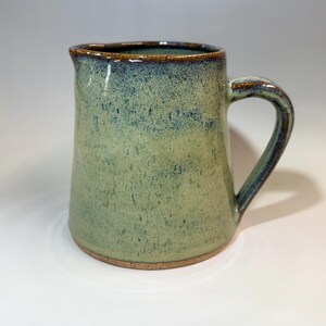 May include: A handmade ceramic pitcher with a handle. The pitcher has a light green glaze with blue and brown accents. The rim and handle are a darker brown. The pitcher is a unique piece of pottery.