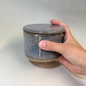 Pu&ograve; includere: Un burriera in ceramica fatta a mano con coperchio. Il piatto &egrave; cilindrico con una glassa blu screziata e una base naturale non smaltata. Il coperchio &egrave; piatto e corrisponde al colore del piatto. La burriera &egrave; tenuta da una mano.
