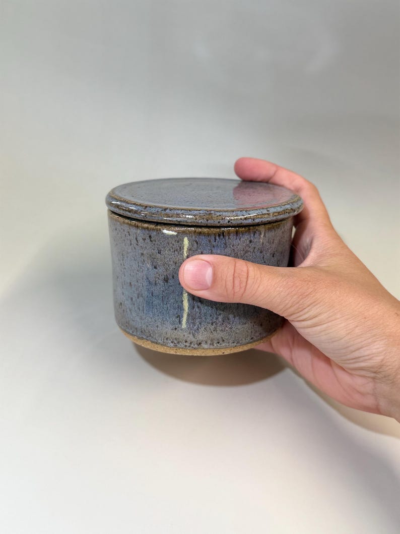 Pu&ograve; includere: Un burriera in ceramica fatta a mano con coperchio. Il piatto &egrave; cilindrico con una glassa blu screziata e una base naturale non smaltata. Il coperchio &egrave; piatto e corrisponde al colore del piatto. La burriera &egrave; tenuta da una mano.