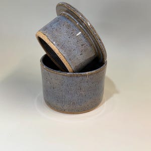 Pu&ograve; includere: Una burriera in ceramica con coperchio. Il piatto e il coperchio sono di colore blu-grigio maculato con un bordo marrone. Il coperchio &egrave; leggermente socchiuso, rivelando l'interno del piatto. La burriera &egrave; di forma cilindrica.