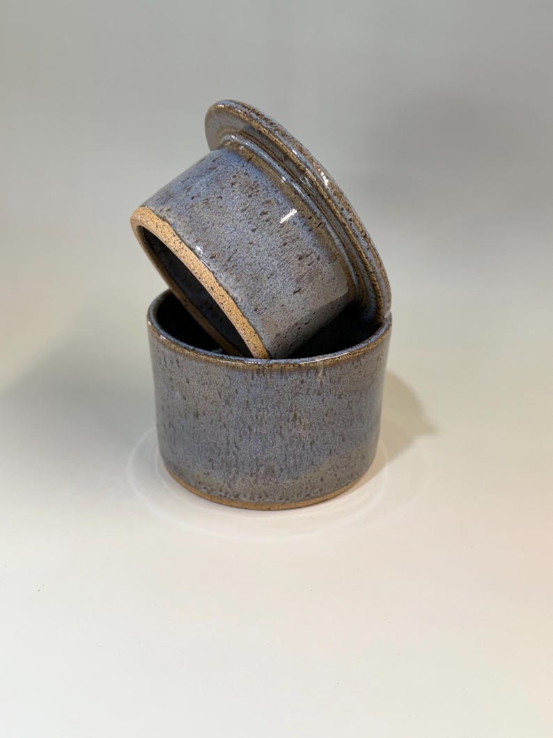 Pu&ograve; includere: Una burriera in ceramica con coperchio. Il piatto e il coperchio sono di colore blu-grigio maculato con un bordo marrone. Il coperchio &egrave; leggermente socchiuso, rivelando l'interno del piatto. La burriera &egrave; di forma cilindrica.