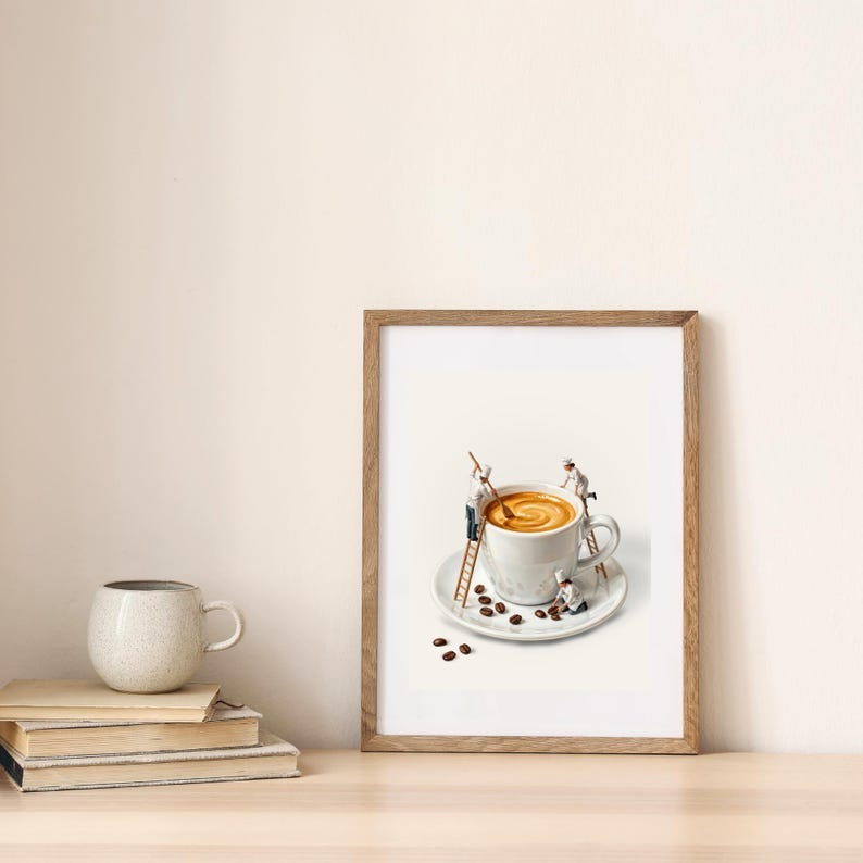 May include: A framed print depicts a whimsical scene of miniature figures working on a large cup of coffee. The coffee cup sits on a saucer with scattered coffee beans. The print is in a wooden frame.