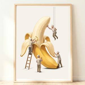May include: A framed art print featuring a large, peeled yellow banana being worked on by four miniature figures dressed as chefs in white uniforms and hats. One figure stands on a wooden ladder, another hangs from ropes, and two stand on the ground.