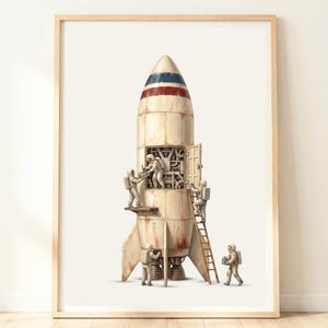 以下が含まれることがあります： クリーム色のロケットに赤と青のストライプが描かれた額入りアートプリント。白い宇宙服を着た4人の宇宙飛行士がロケットの周りで作業しており、1人がはしごを登り、他のメンバーがロケットの外側で作業している様子が描かれています。
