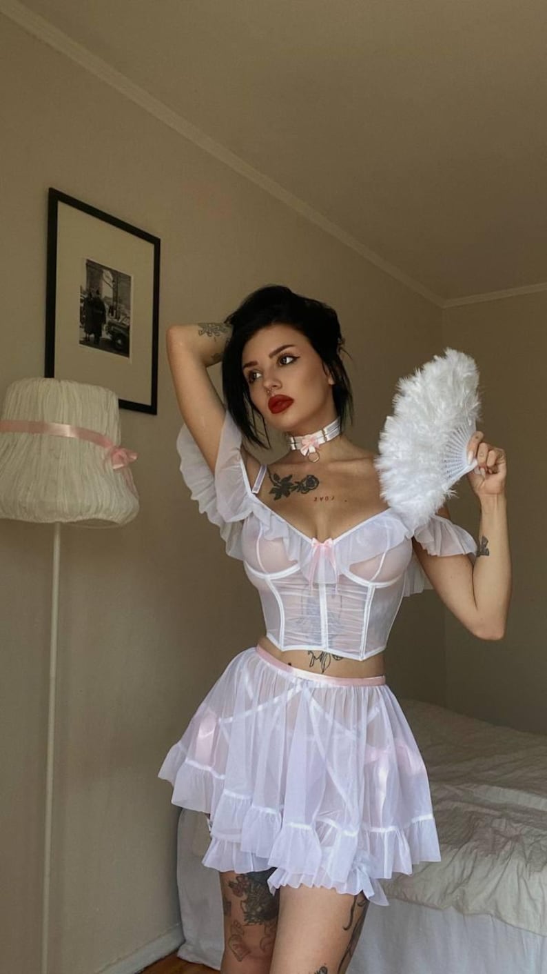 May include: A white lingerie set featuring a sheer corset top with ruffled sleeves and a matching skirt with pink trim. A white feather fan is held. The outfit has a delicate, feminine design.