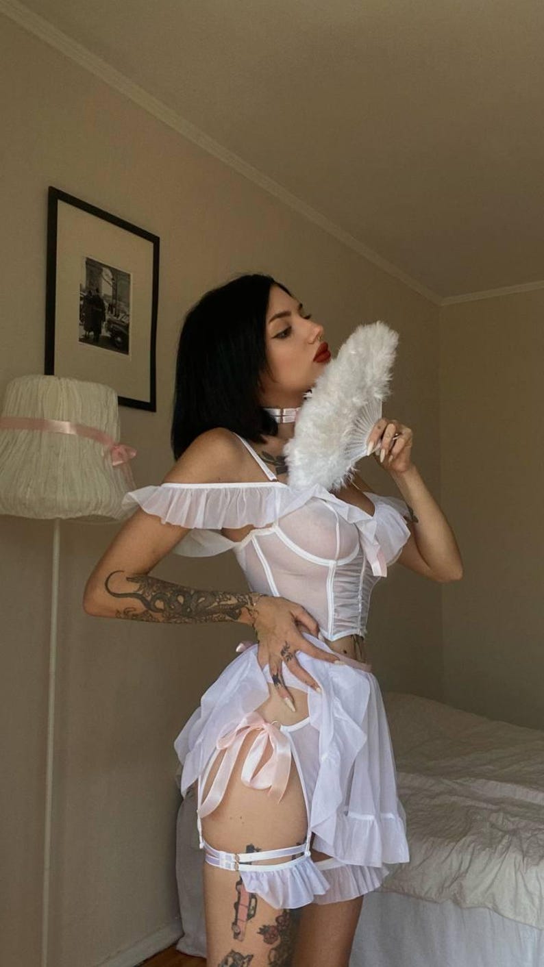 May include: White lingerie set with a sheer corset top, ruffle sleeves, and a matching skirt. The set includes a garter belt with pink bows. A white feather fan is held up. The outfit is designed with delicate details.