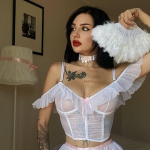 May include: White lingerie set with a sheer corset top, ruffle sleeves, and a matching skirt. The set features a pink ribbon bow. A white feather fan is held in the hand.