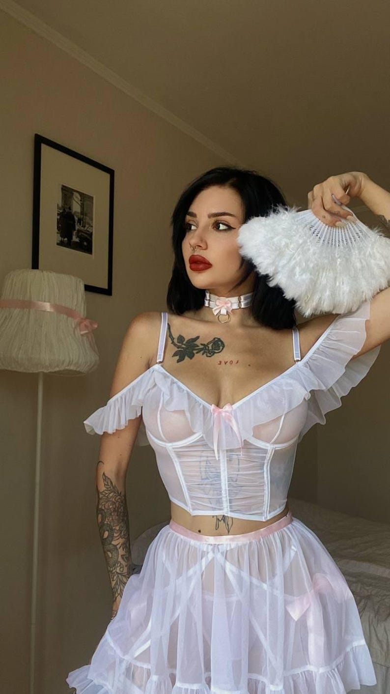 May include: White lingerie set with a sheer corset top, ruffle sleeves, and a matching skirt. The set features a pink ribbon bow. A white feather fan is held in the hand.