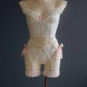 May include: A white lingerie set, comprising a bustier, high-waisted shorts with suspender straps, and a choker, displayed on a mannequin. The bustier, shorts, and choker are adorned with pink bows. The background is a dark blue colour.