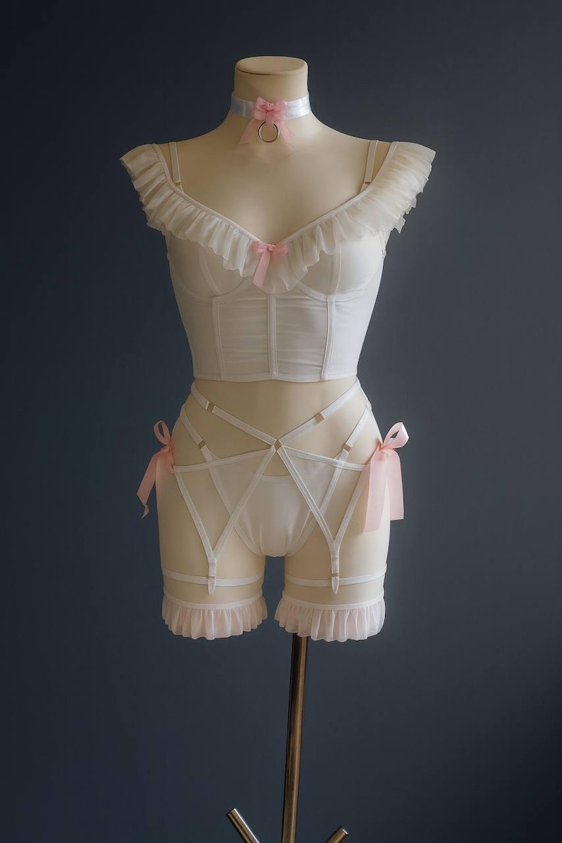 May include: A white lingerie set, comprising a bustier, high-waisted shorts with suspender straps, and a choker, displayed on a mannequin. The bustier, shorts, and choker are adorned with pink bows. The background is a dark blue colour.