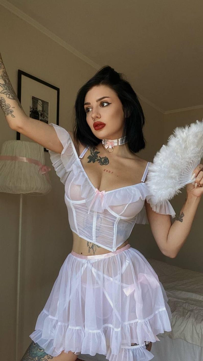 May include: A white lingerie set with a sheer corset top, ruffle sleeves, and a matching skirt, all with pink bow details. A white feather fan is held in the hand.