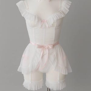 May include: White lingerie set on a mannequin, featuring a ruffled corset, matching shorts with garter straps, and a choker. Delicate pink bows accent the corset, choker, and shorts. The set is made of sheer fabric.