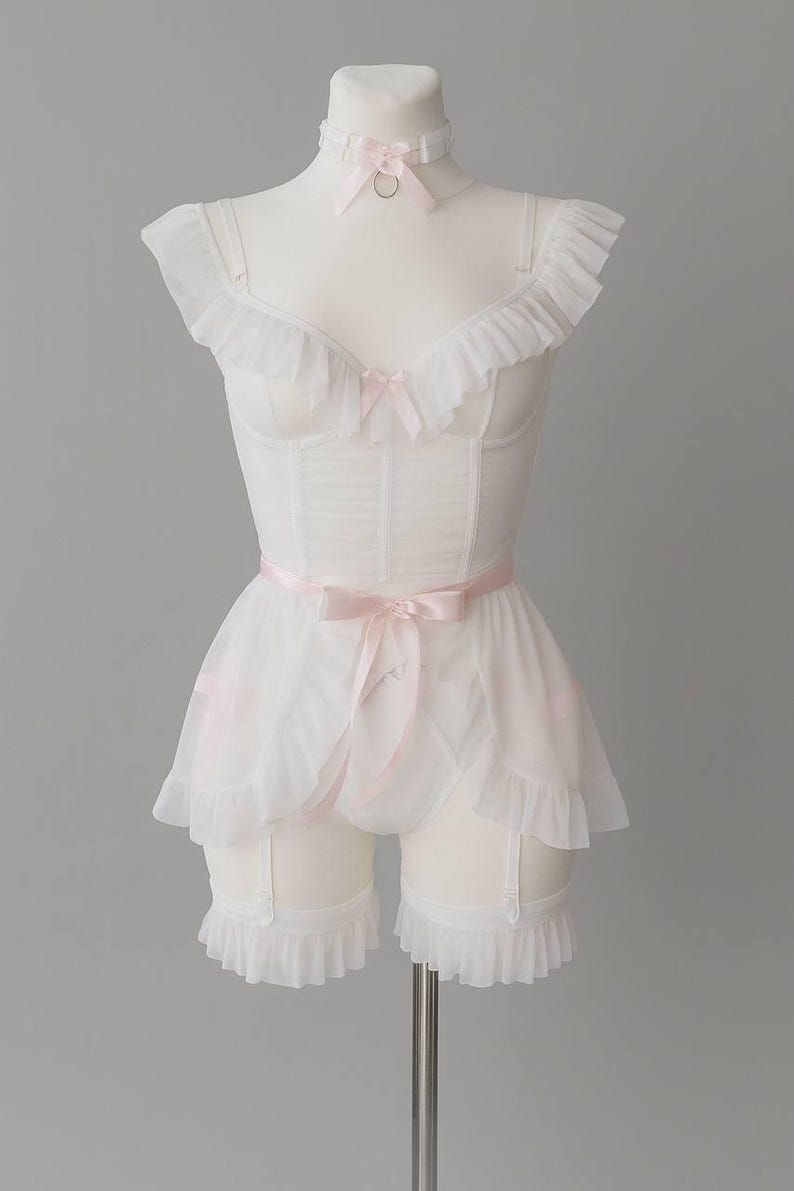 May include: A white lingerie set displayed on a mannequin. It includes a corset with ruffles, matching shorts with suspender straps, and a choker. Pink bows adorn the corset, choker, and shorts. The set is made of sheer fabric.