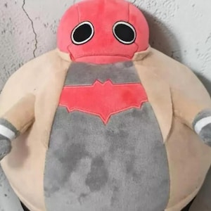 Peut inclure: Une peluche ronde avec une tête rouge et deux grands yeux circulaires noirs. Le corps est beige avec un panneau central gris affichant un emblème rouge en forme de chauve-souris. Les bras sont beiges avec des mains grises et des bandes blanches.