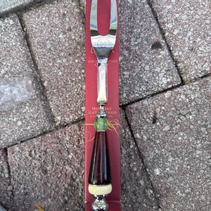May include: A carving fork with a stainless steel head and a decorative handle. The handle features a dark brown cone shape, a cream-colored band, a green bead, and a silver base. The fork is on a red box with the text "HOLIDAY GATHERINGS".