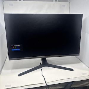 Puede incluir: Un monitor de ordenador Samsung negro con un bisel delgado y un soporte negro. La pantalla muestra un mensaje que dice "Check Signal Cable". El monitor está sobre una superficie blanca.
